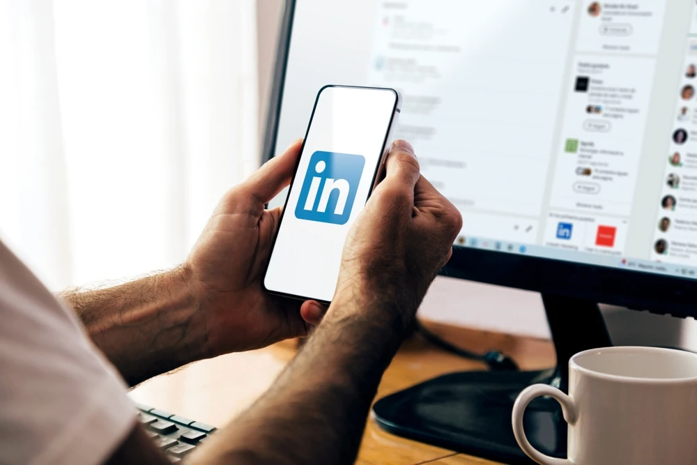 Smartphone met LinkedIn-app, werkplek met computer en koffiemok op achtergrond.