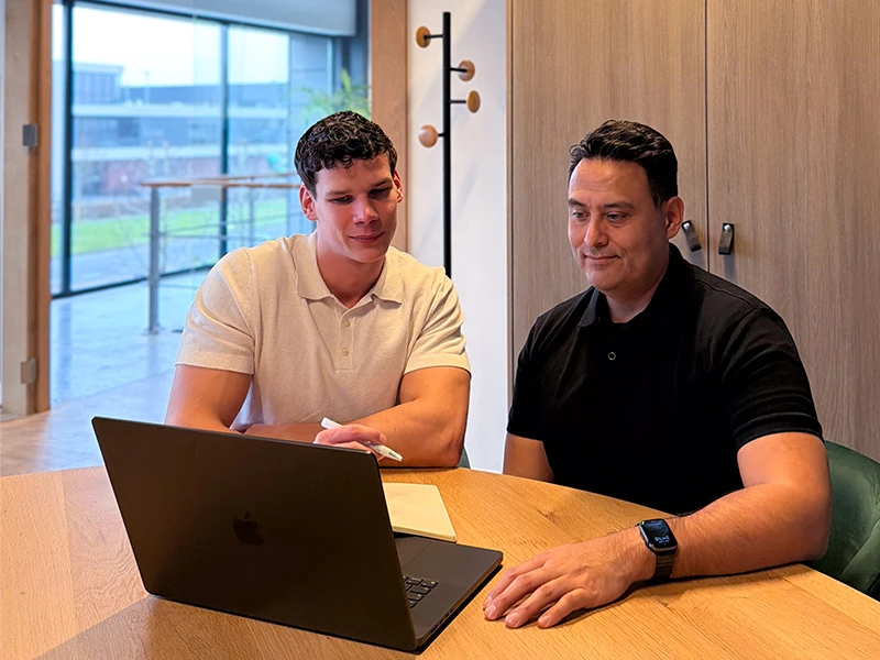 David en Max zitten aan tafel en bespreken samen strategie op een laptop.